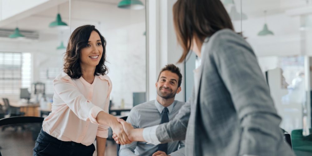 Smiling young businesswoman shaking hands with an office colleague Smiling young businesswoman shaking hands with a coworker during a meeting with colleagues around a table in an office boardroom